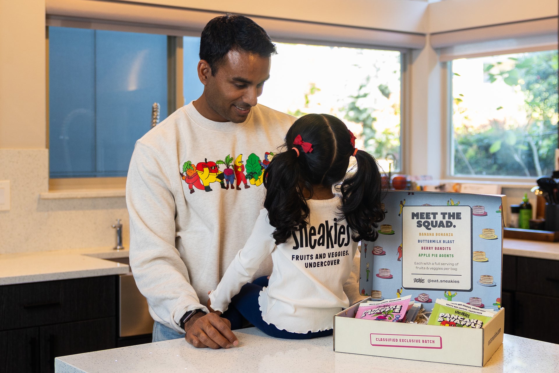 Arun, founder of Sneakies, at his kitchen counter with his young daughter and an open box of Sneakies pancake mixes