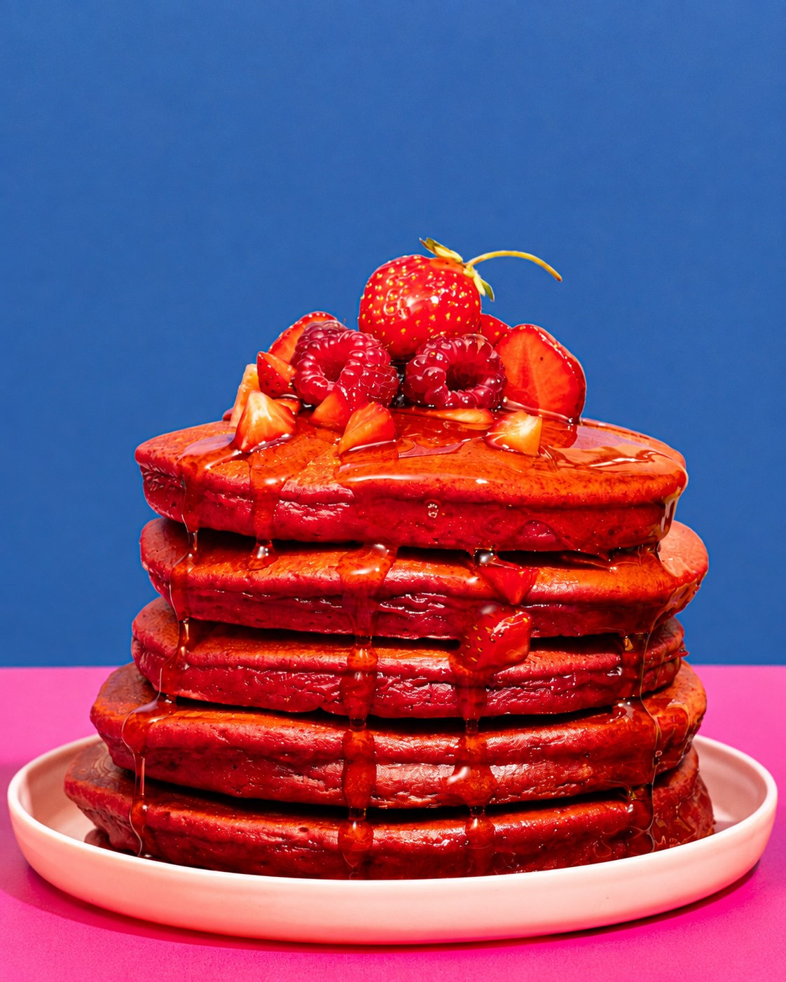 A stack of five Berry Bandits pancakes on a plate, topped with strawberries and raspberries and covered in maple syrup.